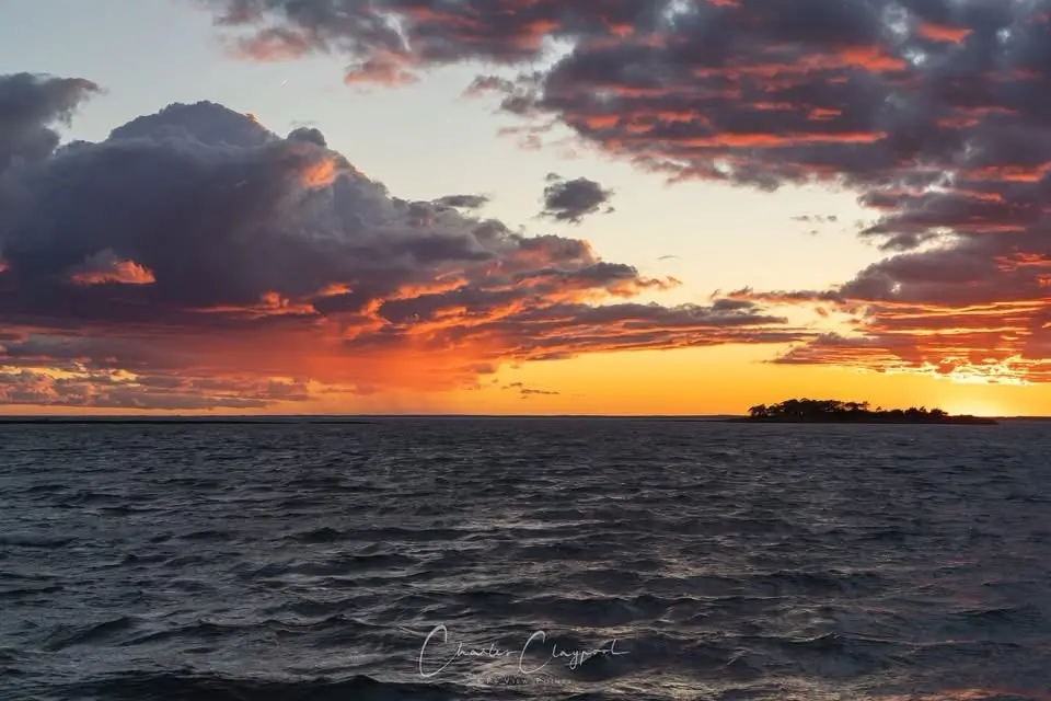 A scenic sunset over a body of water with dramatic orange and purple clouds, a distant island silhouette, and rippling…