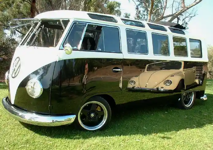 A black-and-white vintage Volkswagen Type 2 van is parked on grass, its shiny side panel reflecting a tan Volkswagen Beetle…