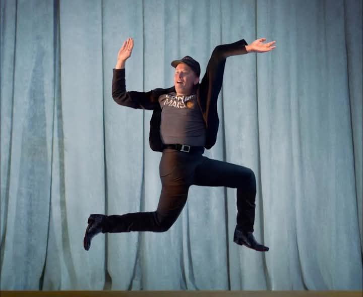 A man in a black jacket, grey t-shirt with 'STAR WARS' text, and a baseball cap is captured mid-air in a dynamic, joyful…