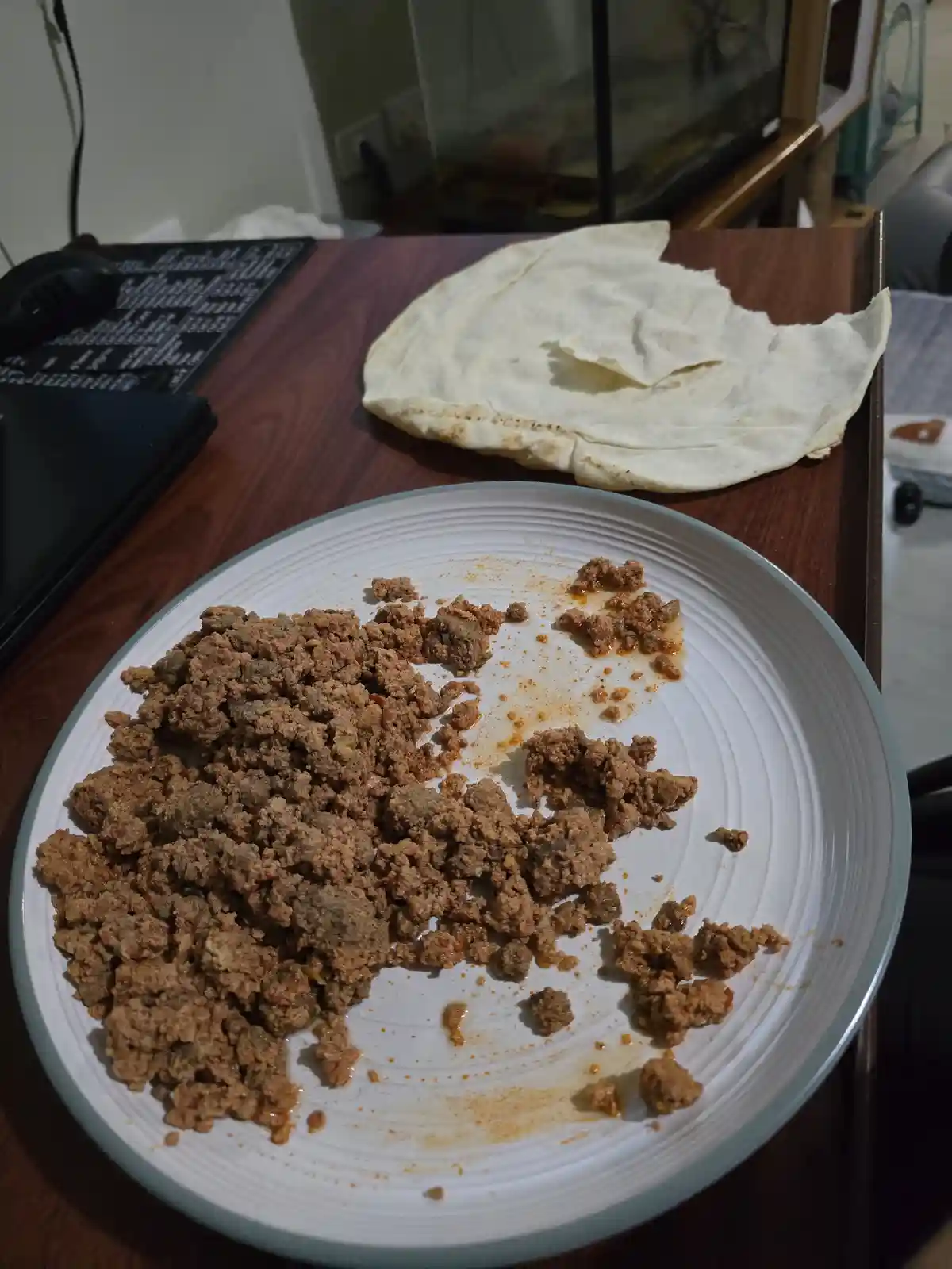 A white plate with mostly eaten ground meat sits on a wooden desk next to a large, untouched tortilla. In the background, a…