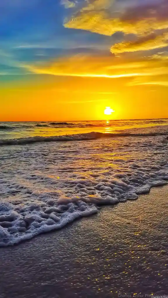 A vibrant beach sunset photo shows golden waves rolling onto dark sand under a sky transitioning from orange to blue, with…
