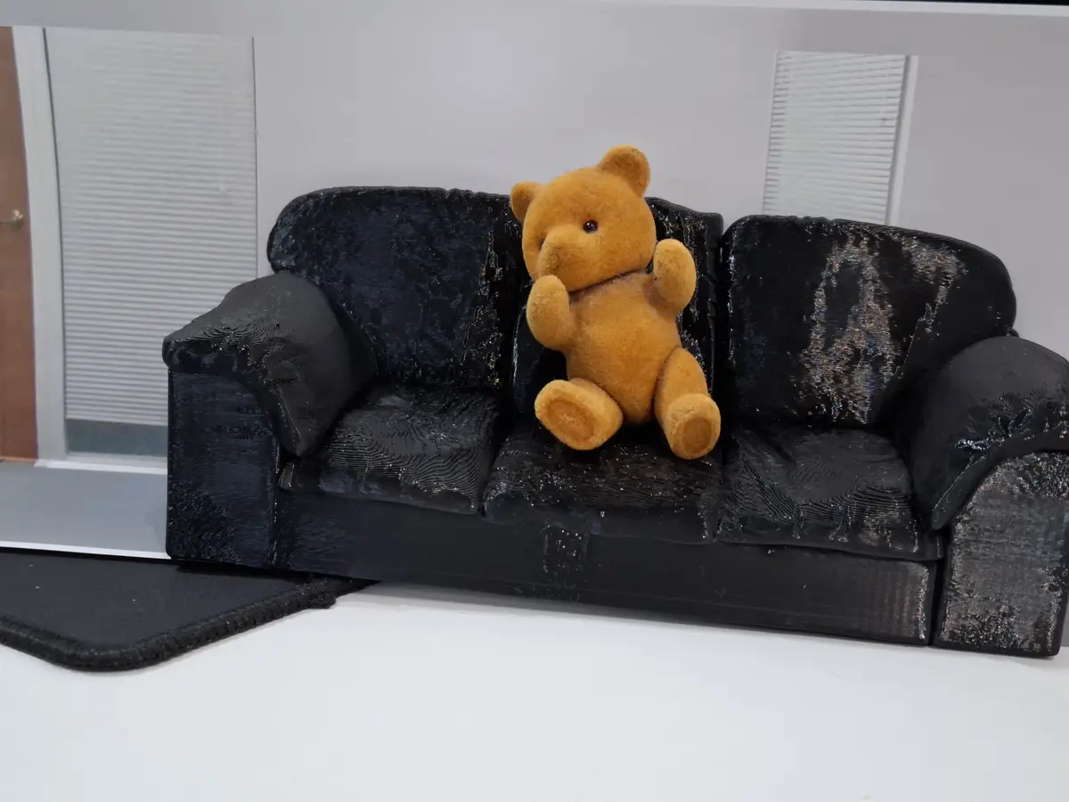 A small, tan stuffed teddy bear sits upright on a miniature black couch that has a visibly rough, 3D-printed texture. The…