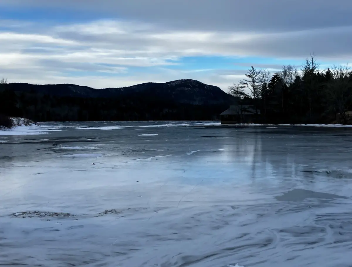 A wide, frozen lake under a cloudy sky, with distant mountains and a small cabin on the far shore. The ice surface is…