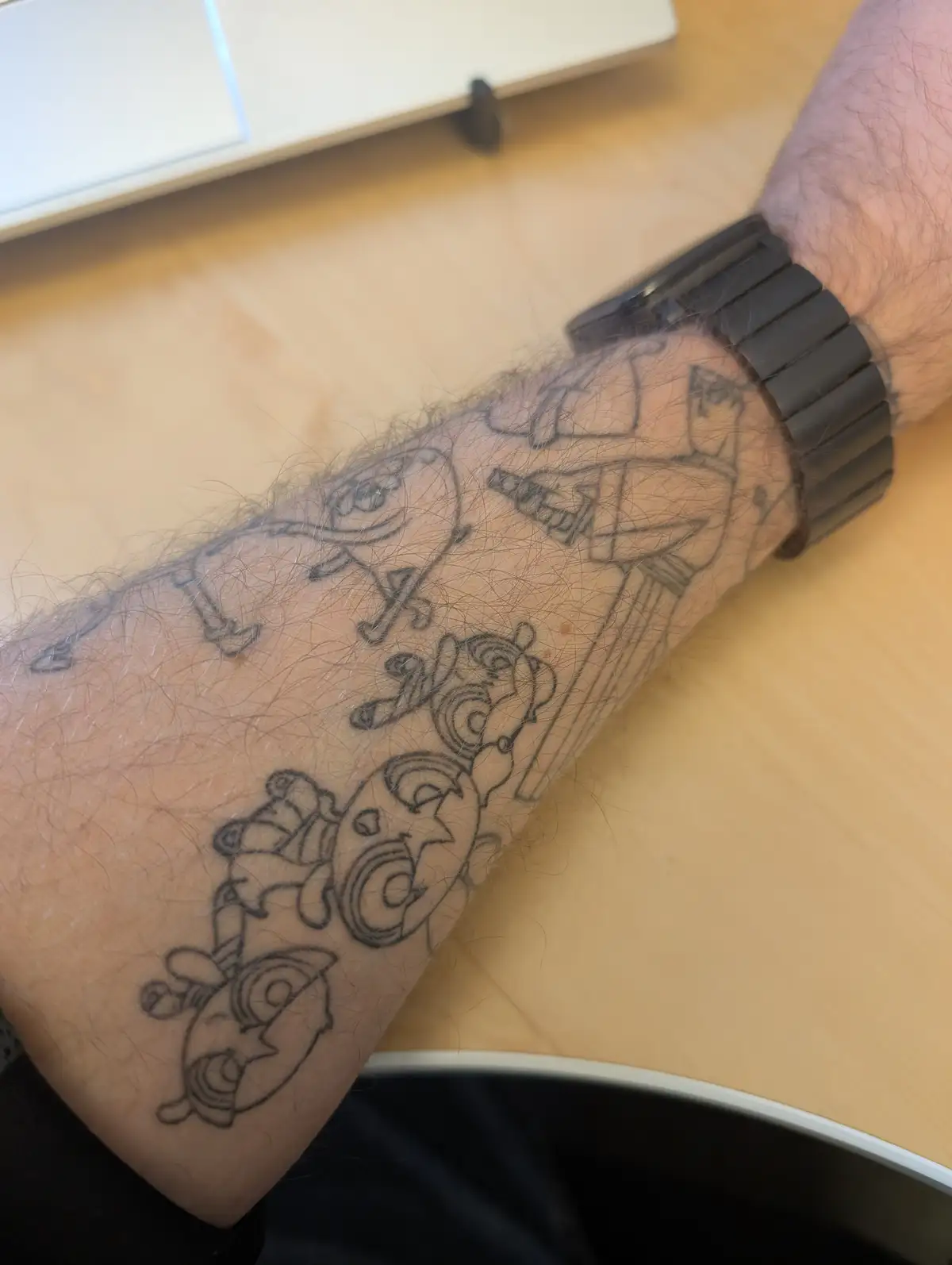A close-up photo shows a person's hairy forearm resting on a wooden desk, displaying a freshly inked black outline tattoo of…