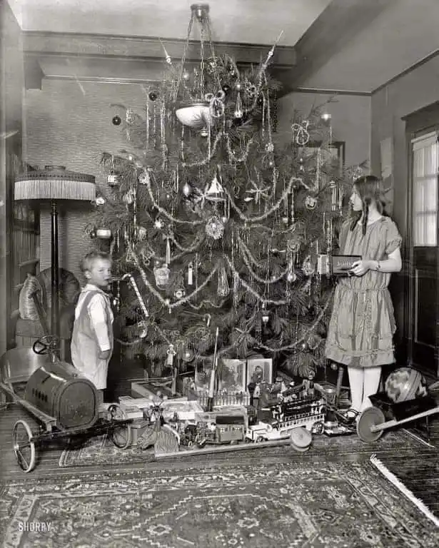 A vintage black-and-white photo shows two children standing beside a heavily decorated Christmas tree. The tree is adorned…