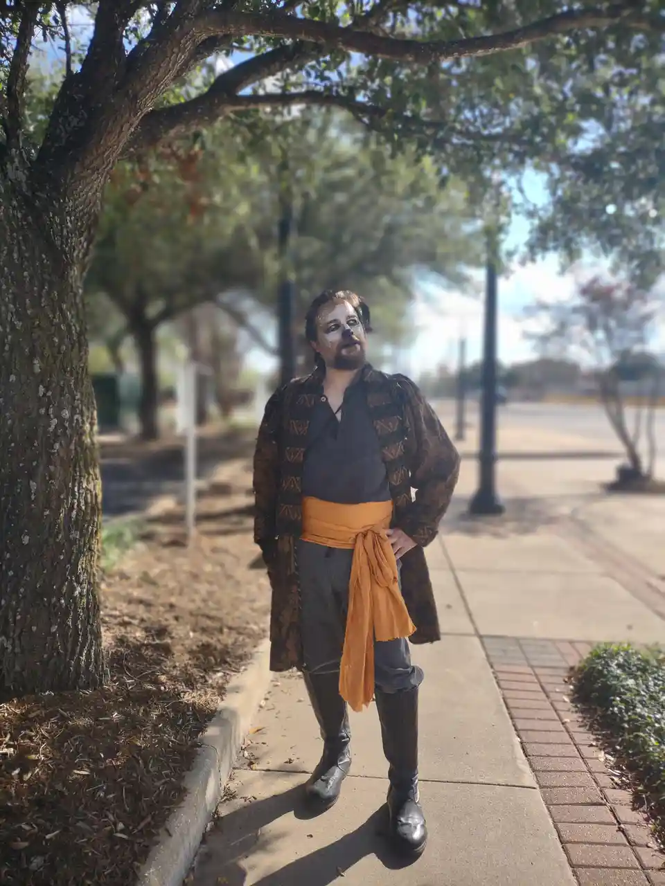 A man dressed in a pirate costume with face paint stands on a sidewalk under a tree. He wears a patterned brown coat, dark…