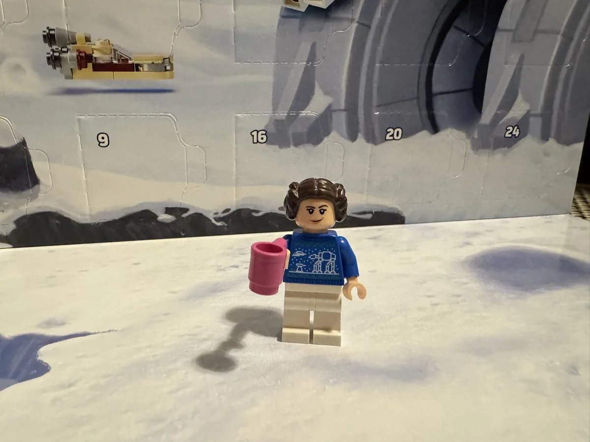 A Lego minifigure of Princess Leia wearing a blue holiday sweater with snowflake and AT-AT designs, holding a pink mug…