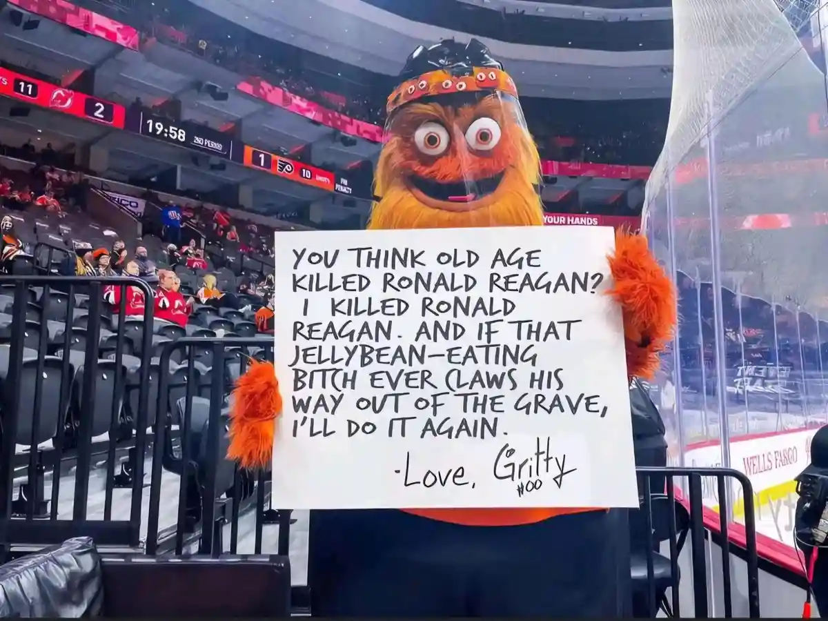 An orange furry mascot named Gritty, wearing a hockey helmet and face shield, holds a handwritten sign at an ice hockey…