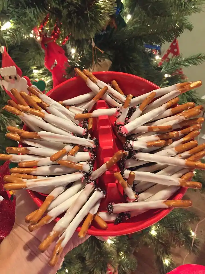 A hand holds a red bowl filled with pretzel rods dipped in white chocolate and sprinkled with black sprinkles, arranged to…