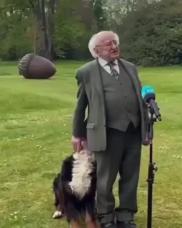 An older man in a grey suit and glasses stands outdoors on grass next to a microphone, holding a dog by its collar. The dog…