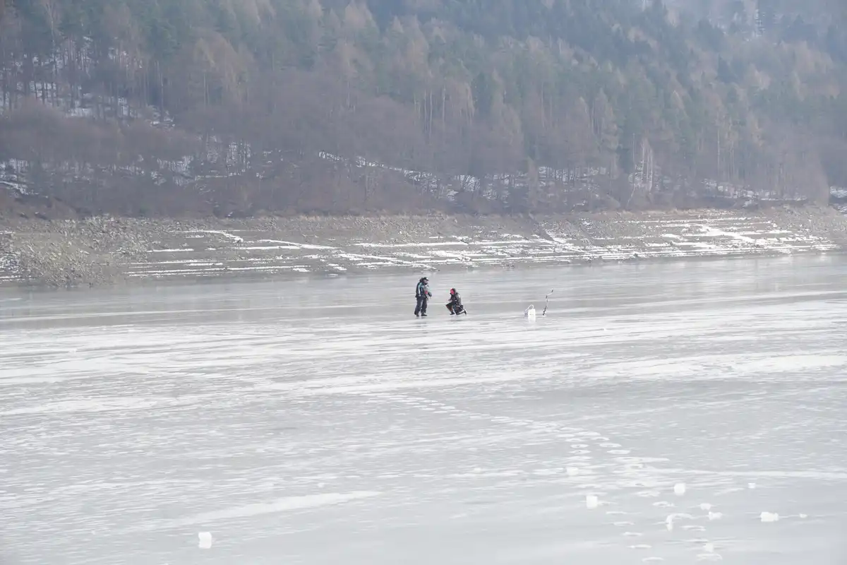 Two people stand on a vast frozen lake, surrounded by snow-dusted forested hills. One person stands upright while the other…
