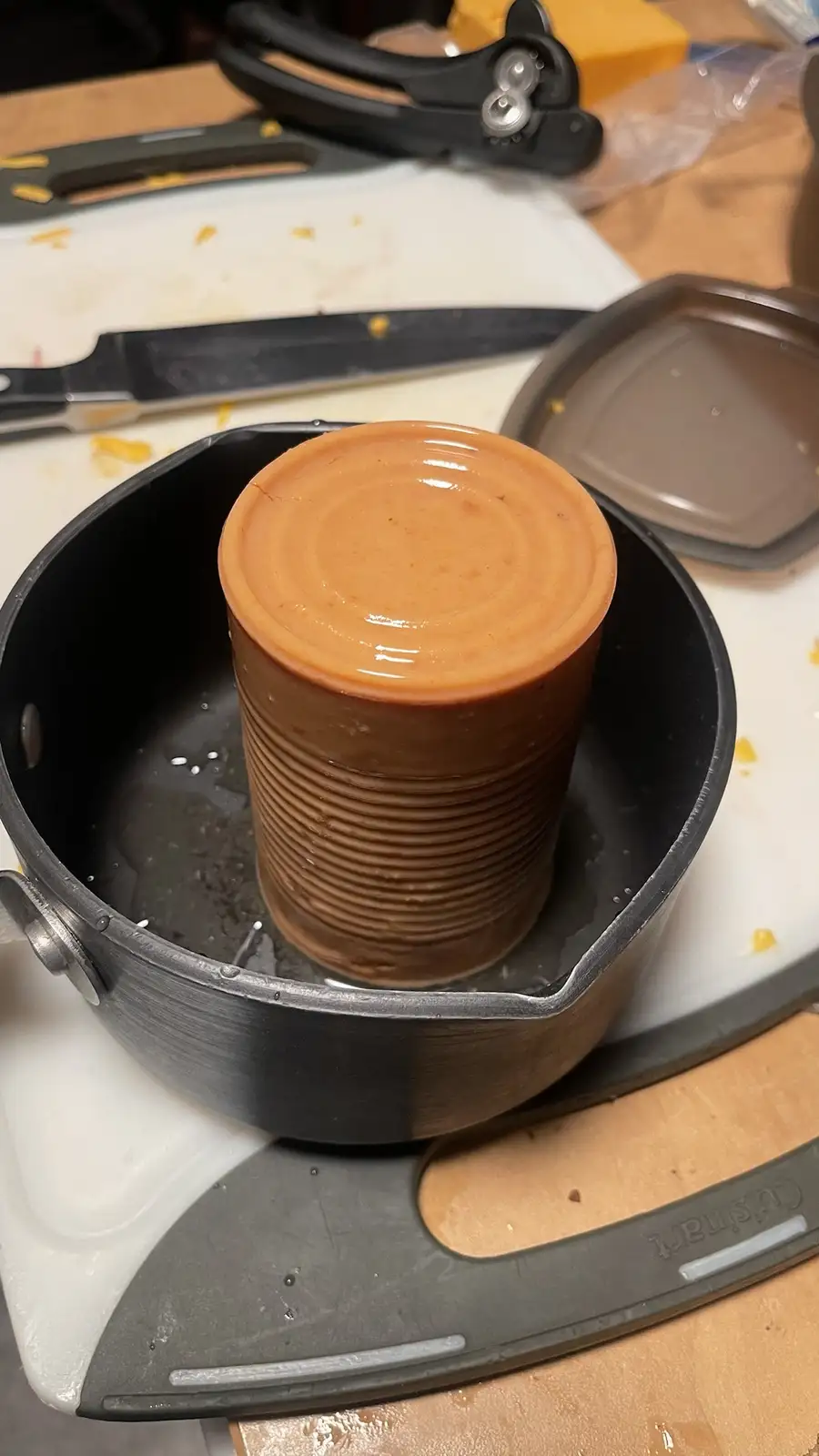 A brown metal can, appearing to be soup, is standing upright inside a small black saucepan filled with water. The saucepan…