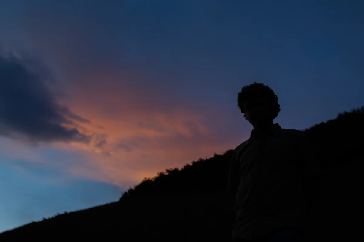 A silhouette of a person stands against a vibrant twilight sky with orange and blue hues, overlooking a dark hillside. The…