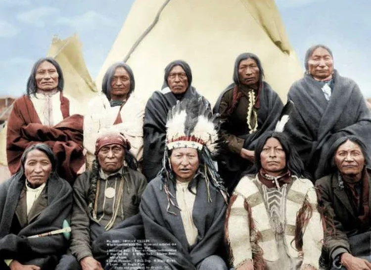 A vintage, colorized photograph shows a group of nine Native American individuals, likely a family, posed formally in front…
