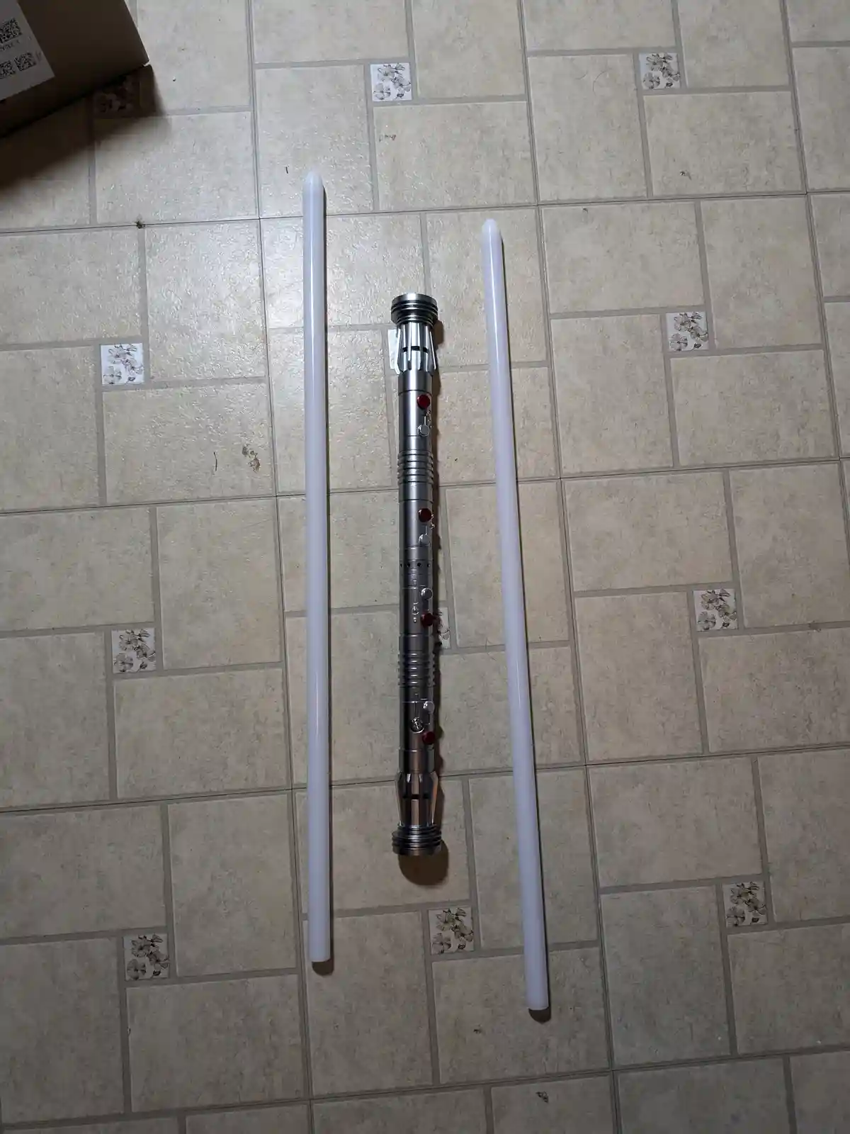 Three white plastic lightsaber hilt components lie on a tiled floor, with two long white cylindrical blades flanking a…