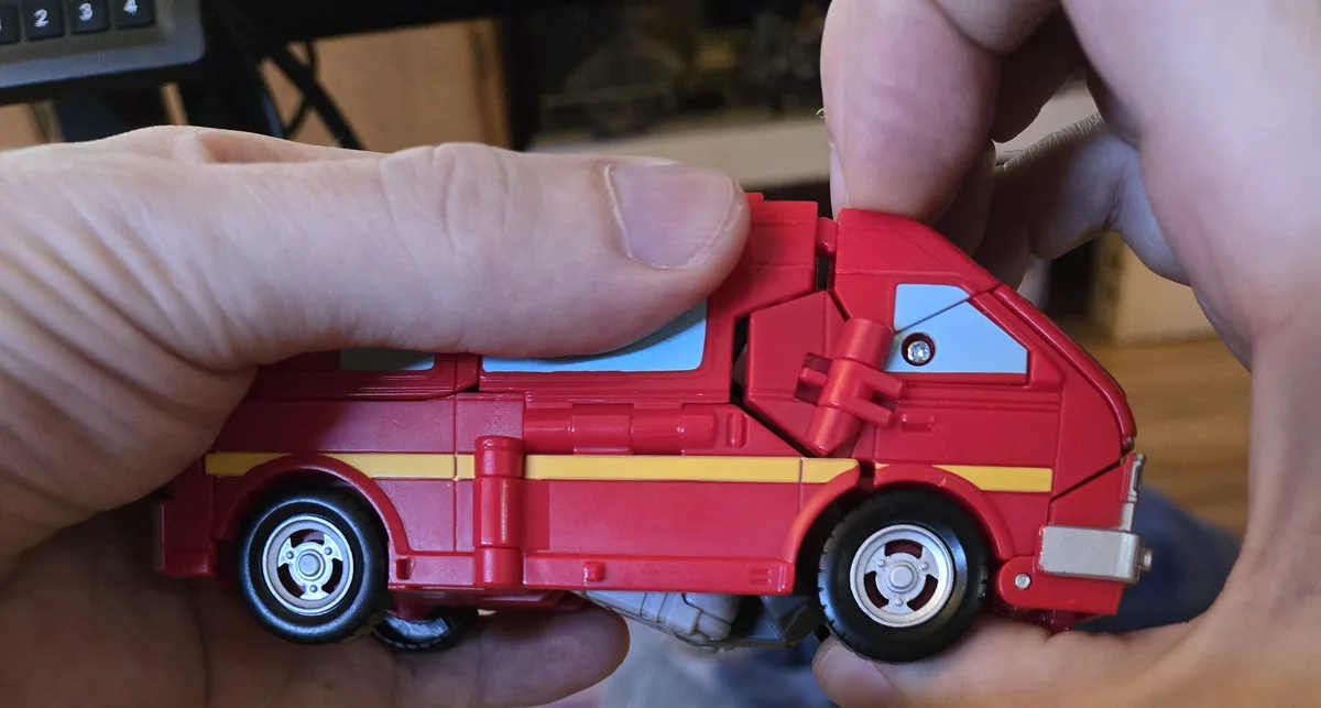 A person holds a red toy vehicle with yellow stripes and black wheels, showing its transforming mechanism. The toy is…