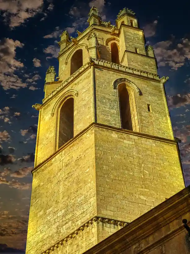 A low-angle photograph shows a tall, ornate stone bell tower at dusk, its surface bathed in warm golden light against a…