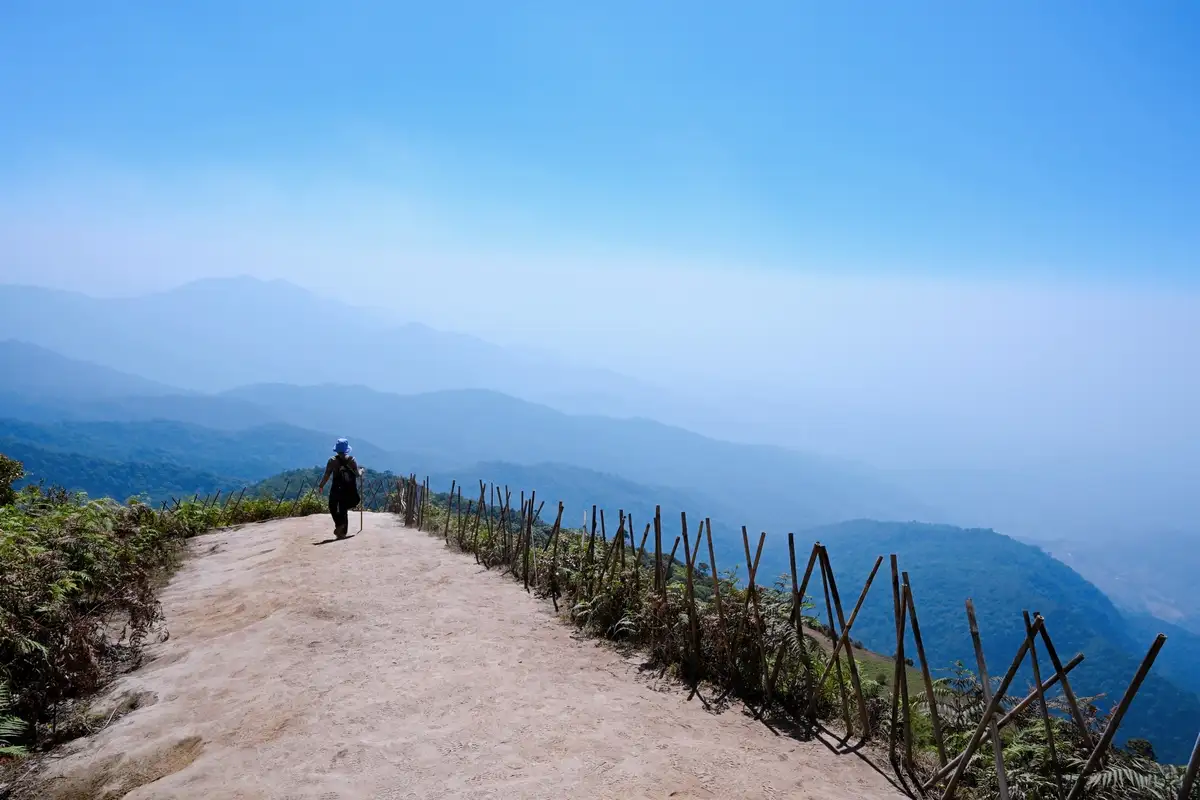A person wearing a blue hat and backpack walks away down a dirt trail on a mountain ridge, with a wooden fence on one side…