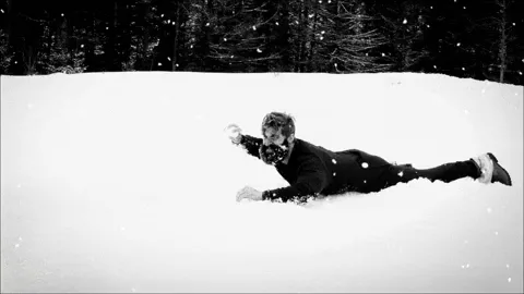 A black-and-white photo sequence shows a man in a dark jacket lying prone in deep snow, throwing a snowball in the top row…
