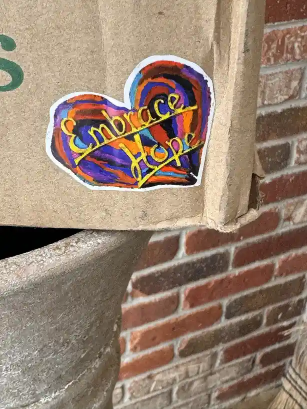A hand-drawn, colorful heart-shaped sticker with swirling rainbow patterns and the words 'Embrace Hope' in yellow cursive is…