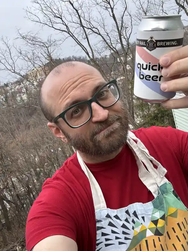 A man with glasses and a beard wearing a red shirt and a colorful apron takes a selfie outdoors holding up a can of 'Rusty…