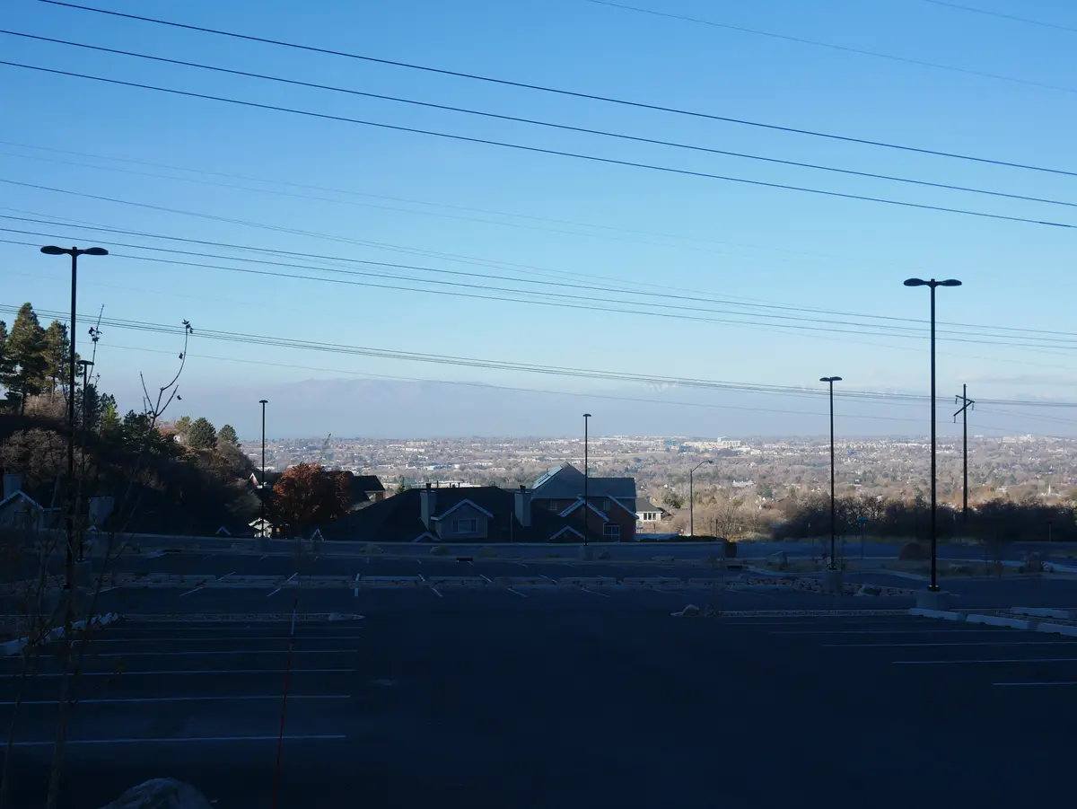 An empty asphalt parking lot with white painted lines, surrounded by streetlights and sparse trees, overlooks a distant…
