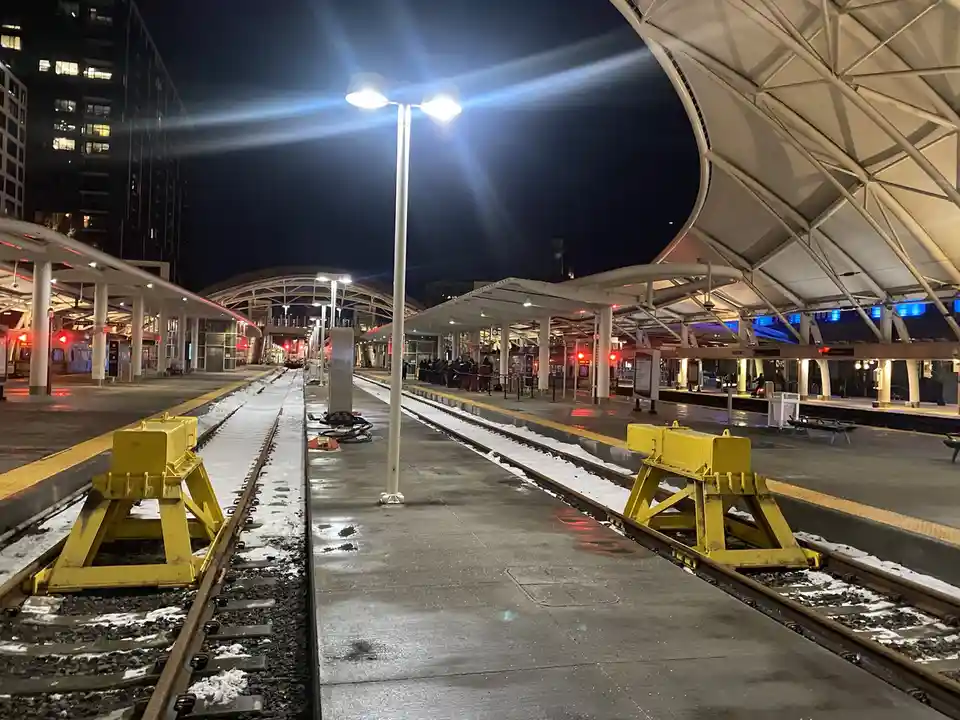 A nighttime photo of an empty train station platform with snow on the tracks and yellow rail stop blocks. A group of people…