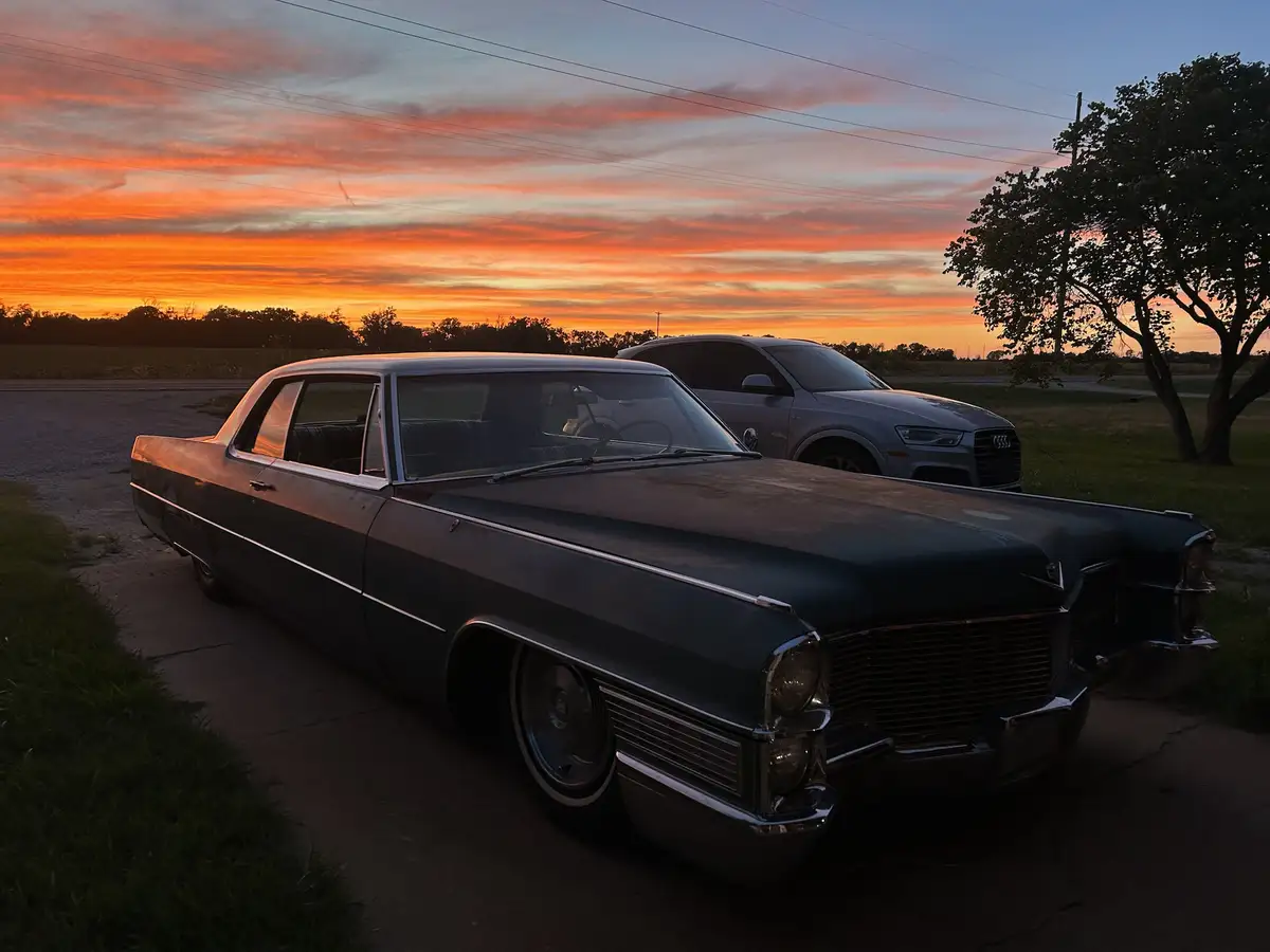 A vintage dark green Cadillac sedan with chrome trim is parked on a driveway at sunset, with a modern silver Audi SUV parked…