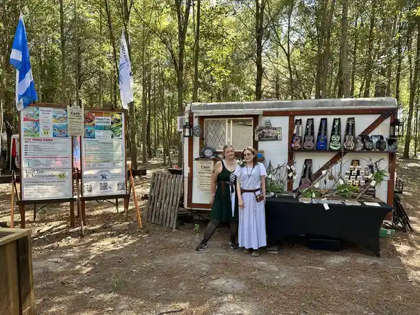 Two women in fantasy-themed dresses stand beside a small trailer set up as a shop in a forest, with a menu board listing…