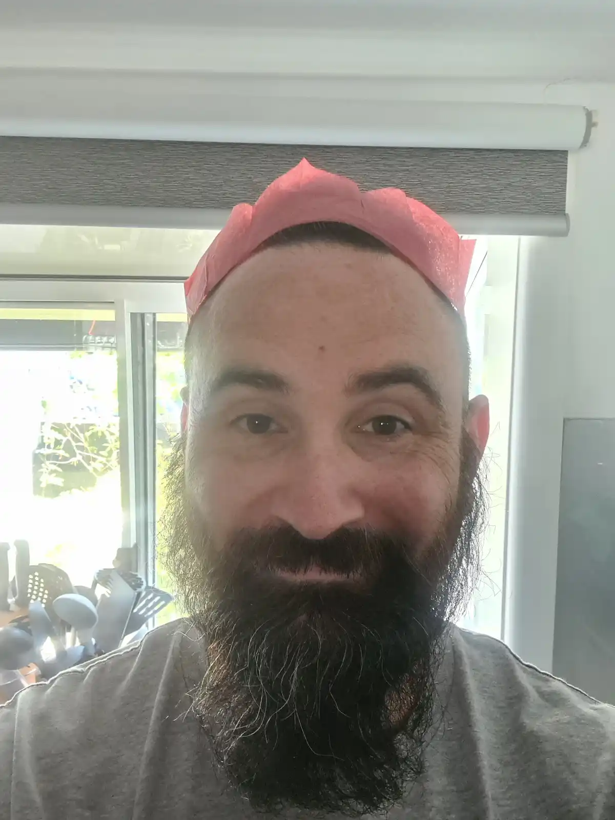 A man with a full beard and shaved head wears a pink paper coffee filter crown while smiling at the camera. He is indoors…