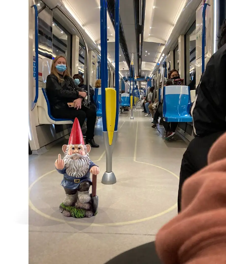 A small garden gnome figurine with a red hat and white beard sits on a subway train floor, holding a shovel and making a…