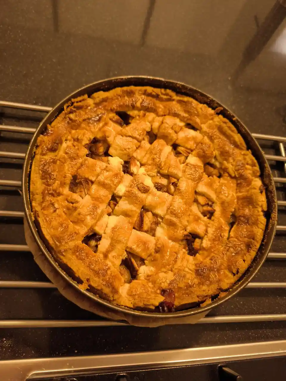 A freshly baked pie with a golden-brown lattice crust sits in a metal baking pan on a cooling rack. The pie appears…