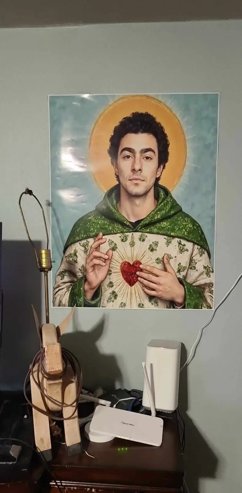 A poster of a man with a halo and a glowing red heart on his chest, styled like a religious saint, is mounted on a wall…
