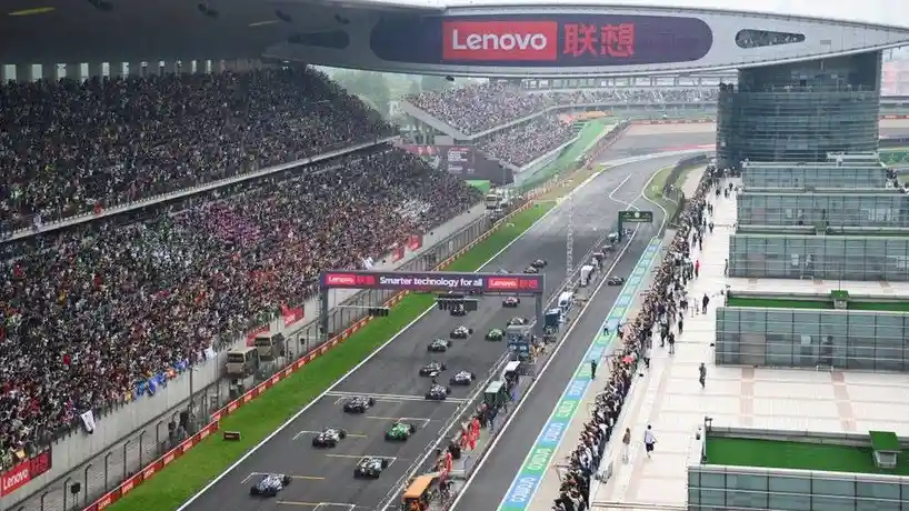 A wide-angle view of a Formula 1 race track filled with spectators in grandstands and along the track, with multiple racing…