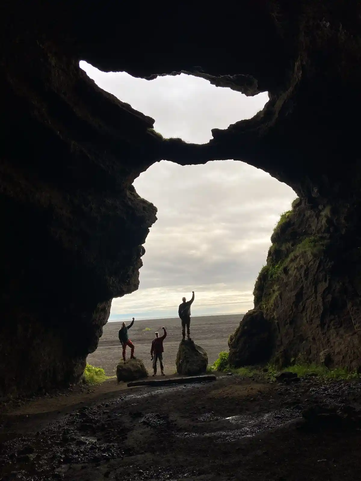 Three people stand inside a large natural rock archway, silhouetted against a cloudy sky, each raising one arm in a…