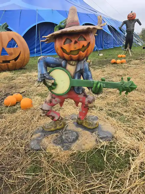 A carved pumpkin scarecrow statue wearing a straw hat, blue jacket, and red pants, playing a green banjo while standing on a…