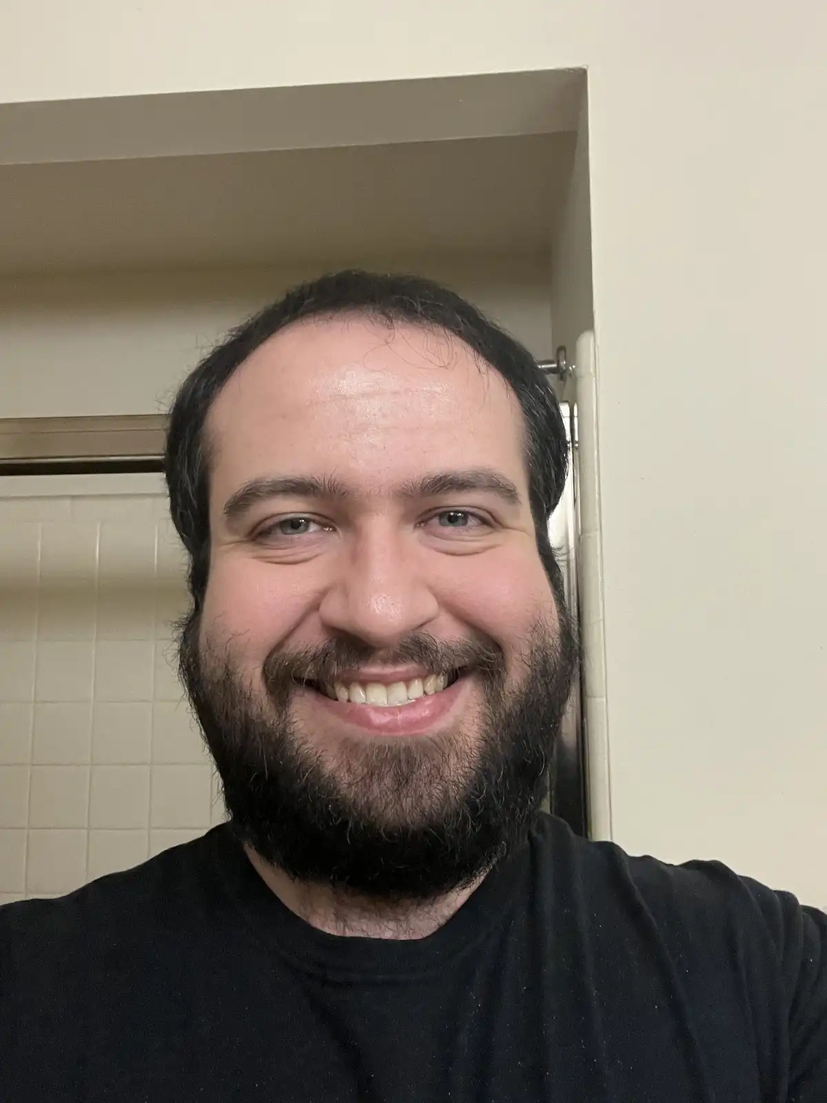 A man with a dark beard and black t-shirt smiles broadly at the camera while standing in a bathroom with a white tiled wall…