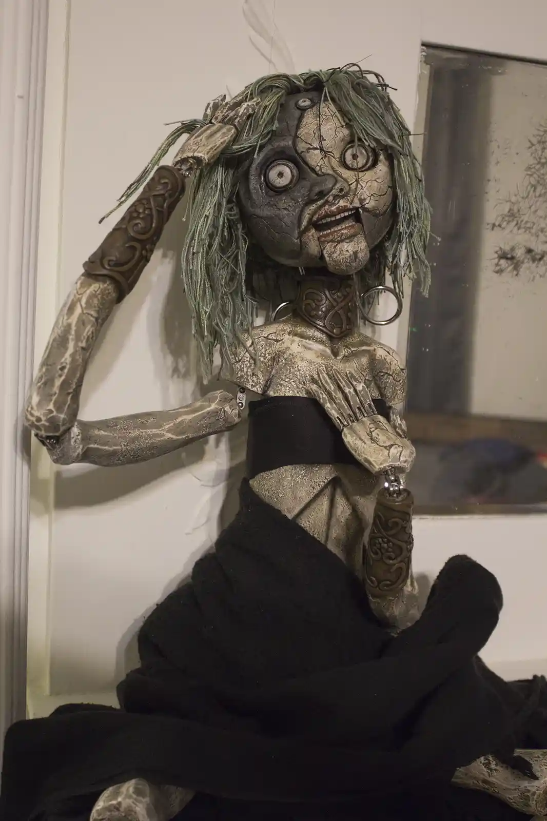 A highly detailed, unsettling doll with cracked, grayish skin, wild green yarn hair, and articulated limbs sits against a…