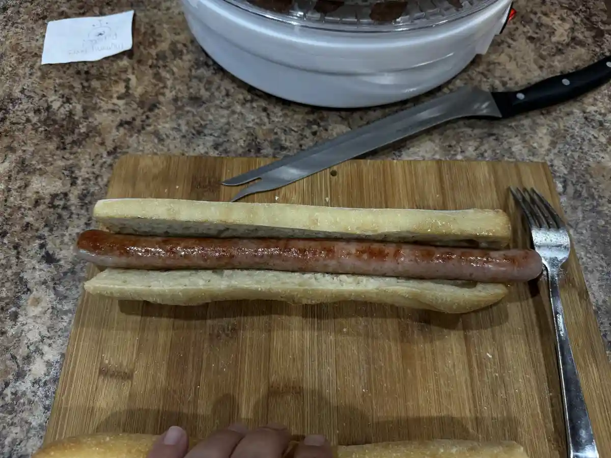 A cooked sausage sits in a long, split bread roll on a wooden cutting board, with a fork and a serrated knife nearby. The…