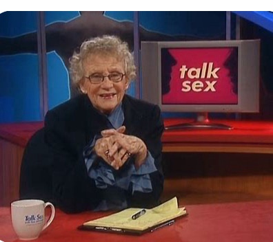 An elderly woman with curly gray hair and glasses sits at a red desk in a TV studio, smiling warmly with her hands clasped…