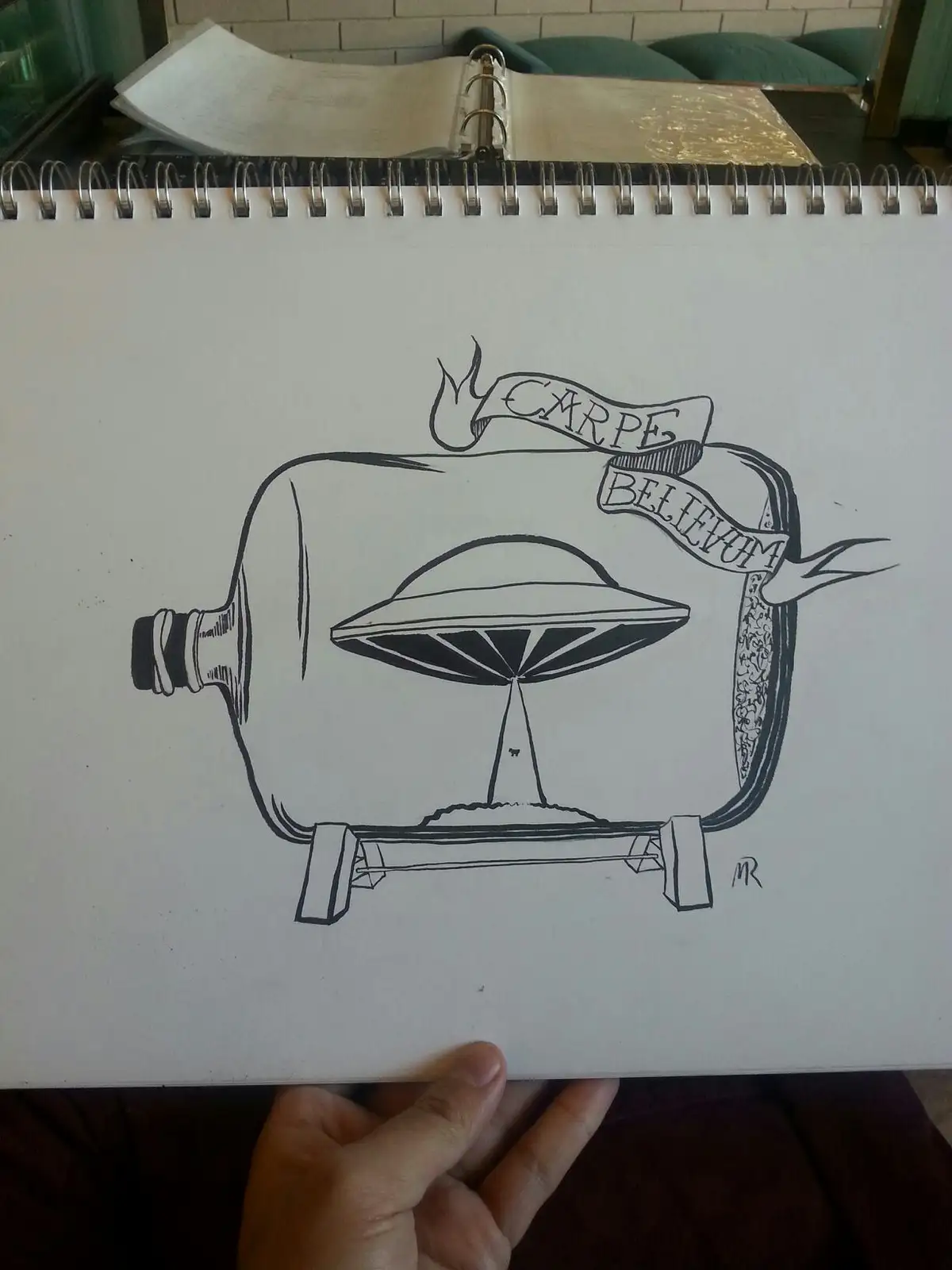 A hand holds a spiral-bound sketchbook open to a black-and-white drawing of a UFO inside a bottle, with a banner above…