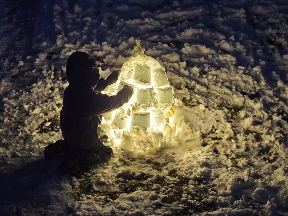 A person is kneeling in a snowy field at night beside a small, illuminated igloo made of blocks of ice. The igloo glows…