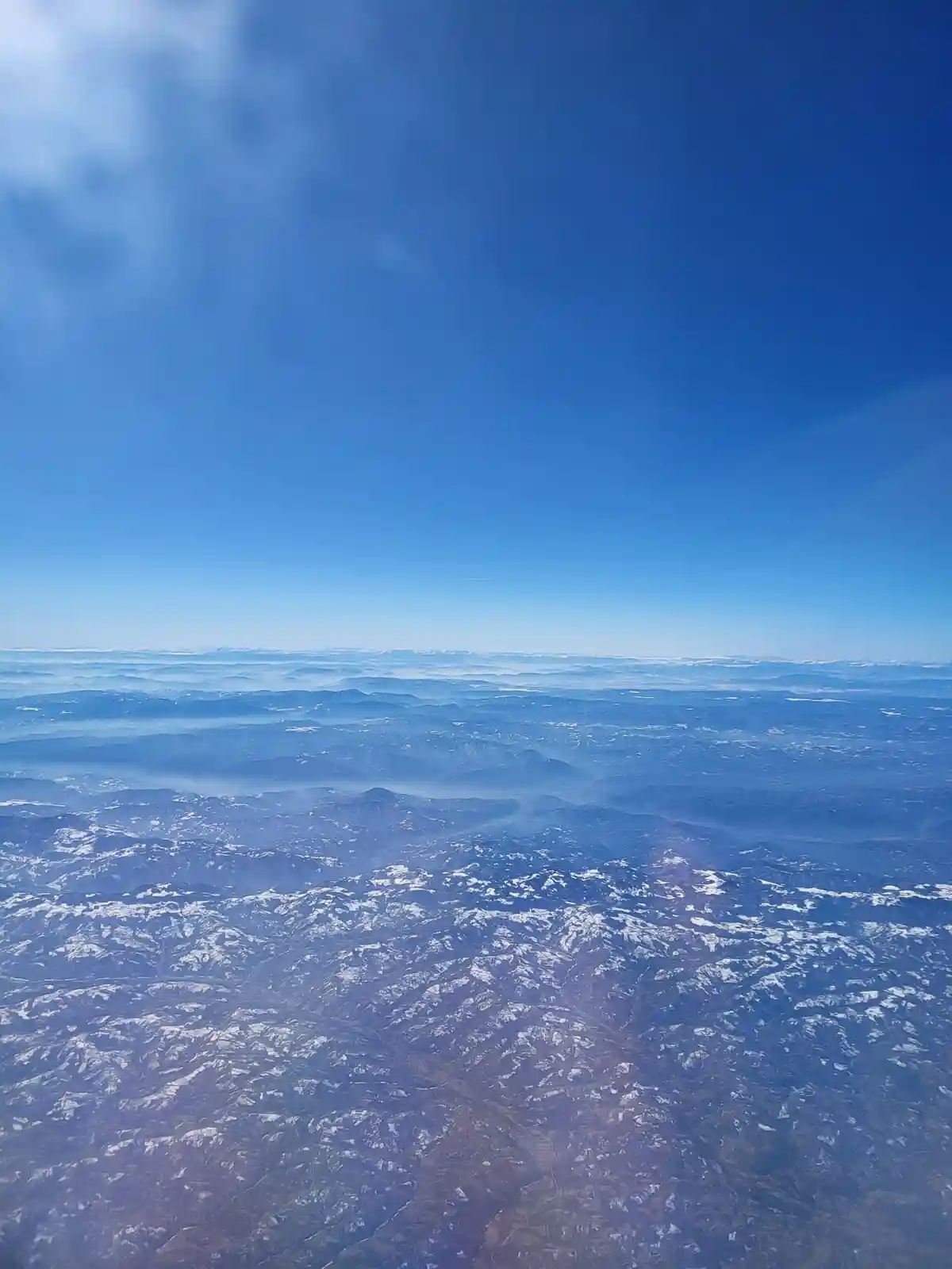 An aerial view from an airplane window shows a vast landscape of rolling mountains partially covered in snow, fading into…