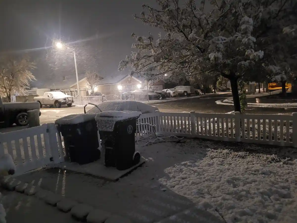 A nighttime photo of a snowy residential street with snow-covered trash bins in the foreground, snow-laden trees, and parked…