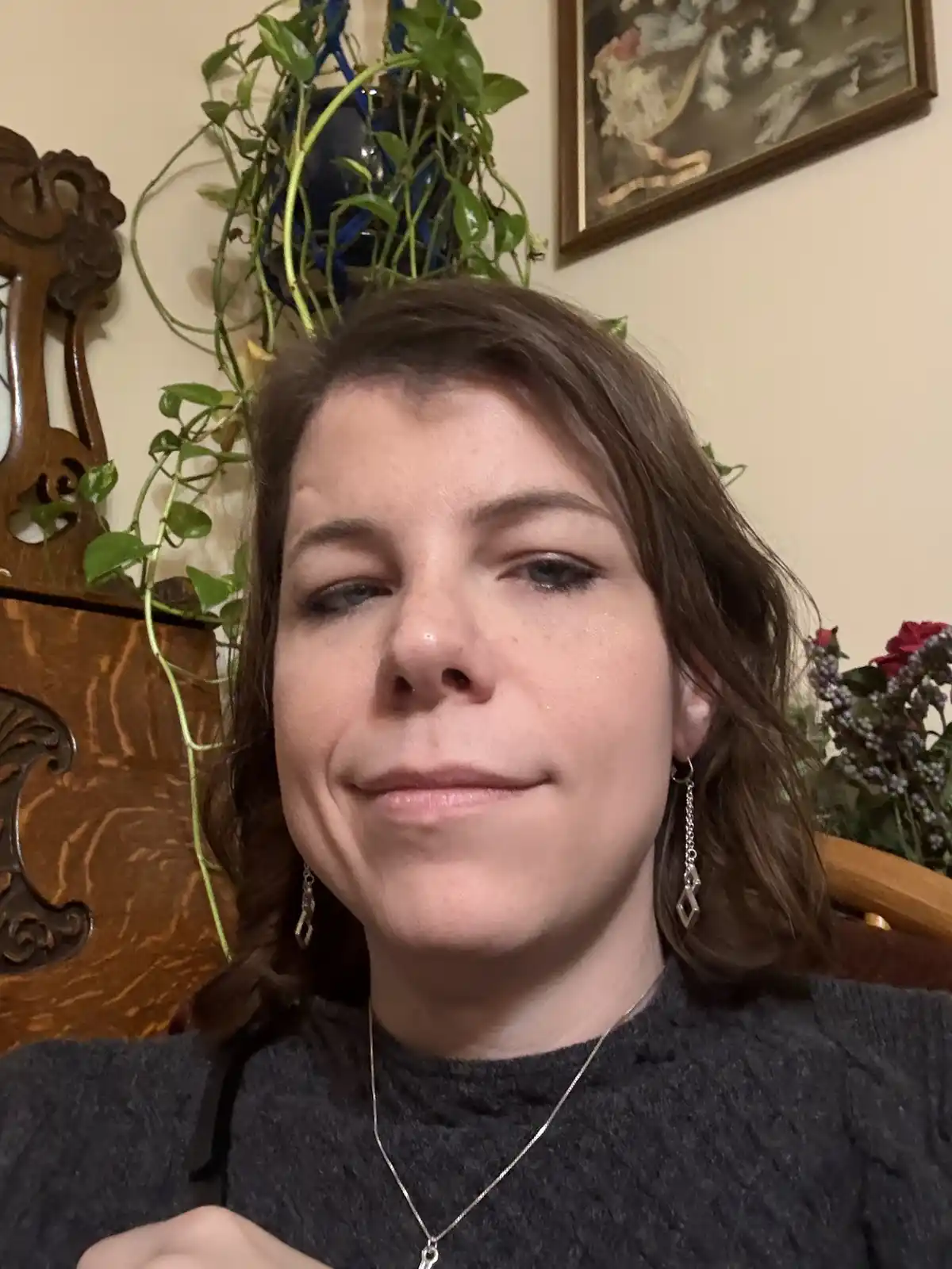 A woman with brown hair and silver earrings looks directly at the camera with a subtle, slightly forced smile while her eyes…