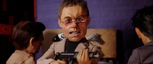 A puppet with a large, exaggerated head and glasses holds a gun while two other puppets face him. The puppet has two pistols…