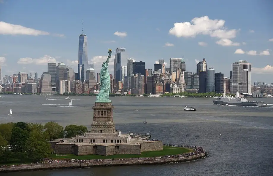 A wide-angle photograph of the Statue of Liberty on Liberty Island, with the New York City skyline and the Freedom Tower…