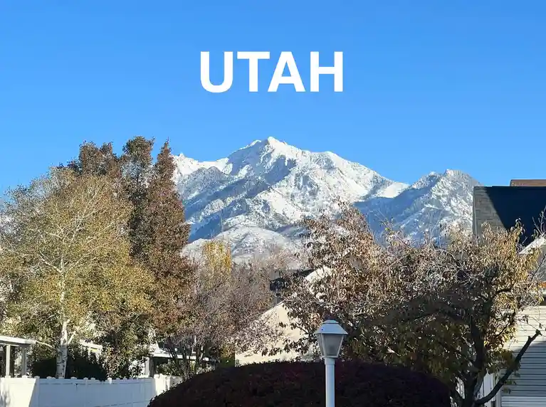 A clear blue sky frames snow-capped mountains visible behind autumn-colored trees and suburban houses, with the word 'UTAH'…