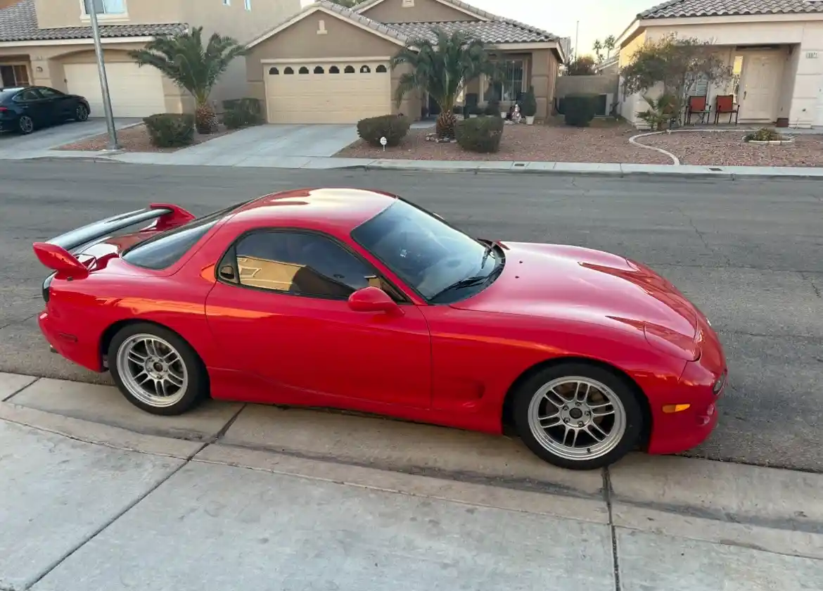 a glossy red mazda rx-7 sports car is parked at an angle on a concrete sidewalk in a suburban neighborhood, with residential…