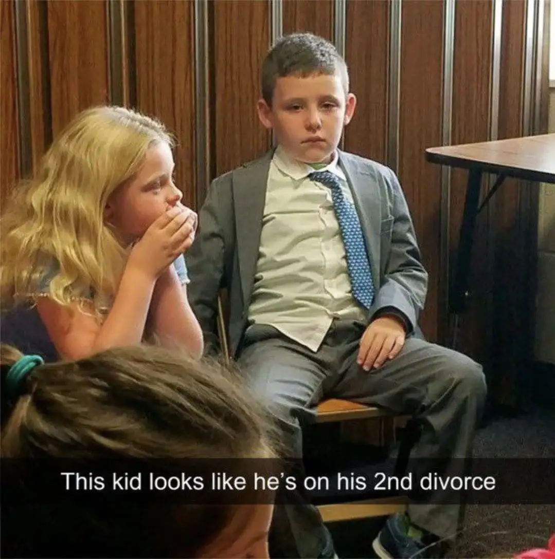 A young boy in a gray suit and blue patterned tie sits with a profoundly bored, unimpressed expression, while a girl beside…